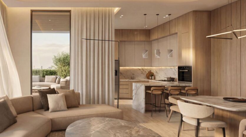Salon et cuisine modernes en espace ouvert à Lunaya Terraces, avec des finitions en bois clair, des touches de marbre, un canapé modulable beige, une table basse ronde, un coin repas et une vue depuis le balcon sur le coin salon extérieur et la verdure à travers de grandes baies vitrées.