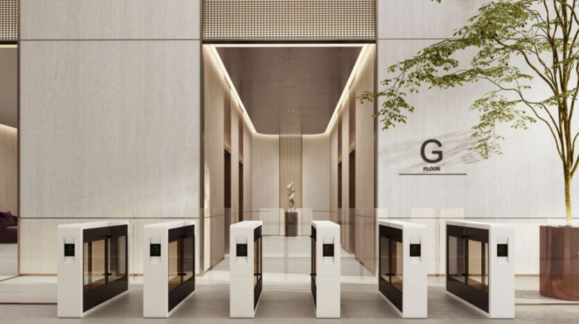Hall d'entrée d'un immeuble moderne équipé de cinq tourniquets électroniques, avec de grands murs beiges, une haute porte d'entrée et un arbre en pot. Une pancarte accrochée au mur indique « G FLOOR », reflétant le style contemporain du quartier O1NE. On aperçoit à l'arrière-plan un couloir où se trouve une statue.