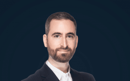 Portrait professionnel d'un homme barbu et cheveux courts, vêtu d'un costume sombre et d'une chemise blanche. Il sourit subtilement et regarde directement la caméra sur un fond bleu foncé, évoquant le