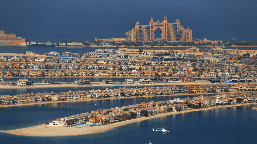 Vista aérea de una ciudad costera que muestra el emblemático Hotel Atlantis en Palm Jumeirah en Dubai, rodeado de lujosas residencias y un mar cristalino, con un helicóptero volando cerca.