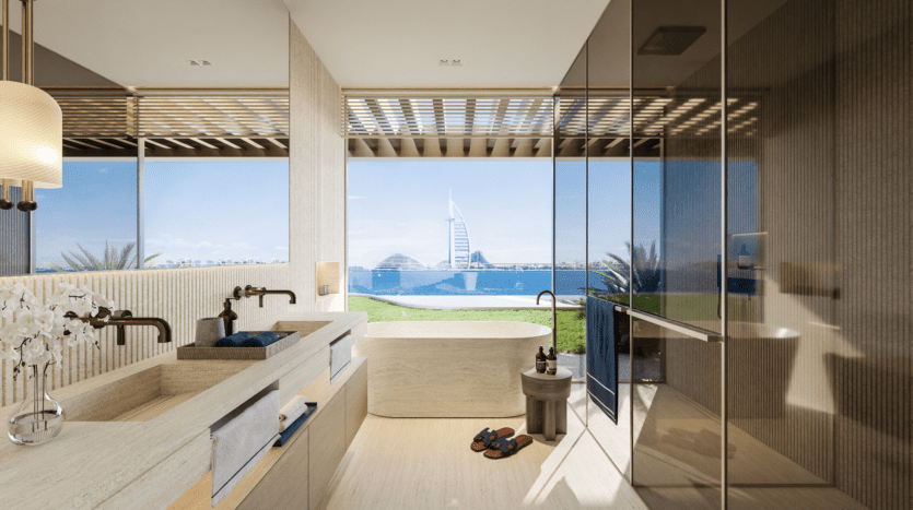 Salle de bains luxueuse avec vue sur un célèbre bâtiment en forme de voile en front de mer sur l'emblématique Palm Jumeirah. Elle dispose de deux lavabos, d'une baignoire îlot et d'une grande fenêtre offrant une vue imprenable sur l'océan et la ville. Une décoration moderne et une lumière naturelle renforcent l'ambiance sereine.