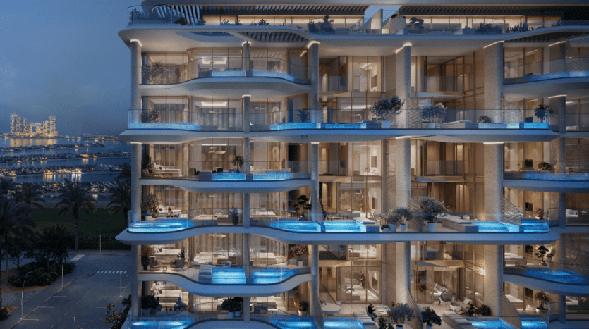 Un bâtiment moderne de plusieurs étages en bord de mer avec des balcons en verre et des piscines privées à chaque niveau. Illuminé la nuit, il offre une vue sur les toits de la ville et sur le Vitalia Palm Jumeirah voisin, bordé de palmiers et d'une route côtière.