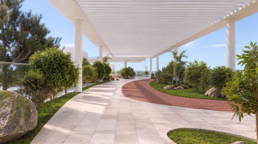 Allée couverte dotée de piliers blancs et d'une pergola moderne au Beverly Crown by HMB, avec un chemin sinueux pavé de dalles beiges et rouges, entouré d'arbres verts, d'arbustes et de rochers, avec en arrière-plan une vue dégagée sur le ciel.
