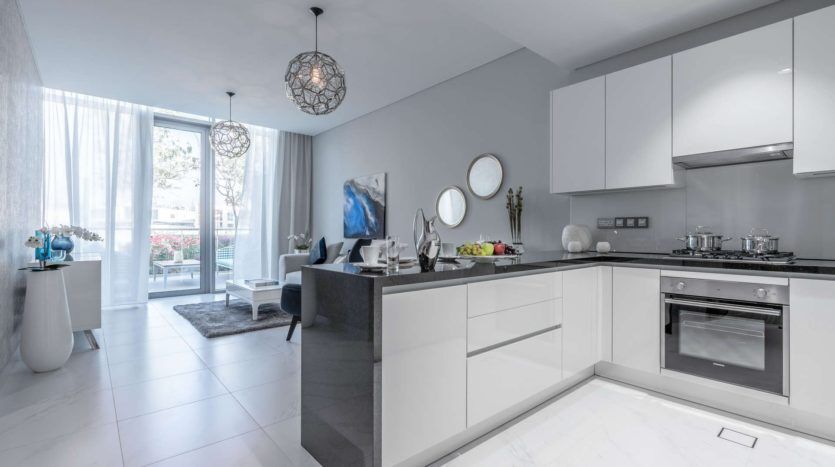 Cuisine et salon modernes et ouverts avec armoires blanches, appareils électroménagers en acier inoxydable et accents bleus. dispose de luminaires artistiques et d'une vue à travers des portes vitrées donnant sur un balcon.