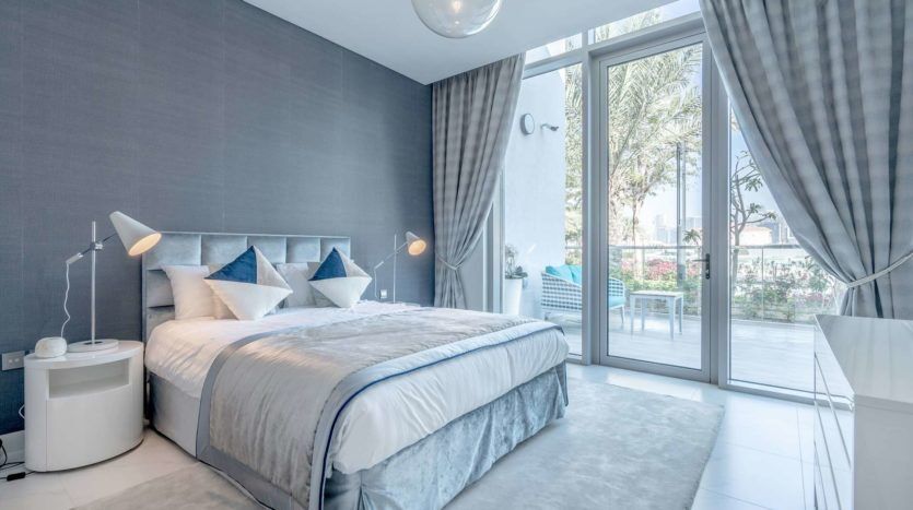Une chambre moderne avec un grand lit, une literie bleue et grise, des murs et des rideaux gris. comprend des tables de nuit avec des lampes et une vue sur un balcon avec de la verdure à l'extérieur.