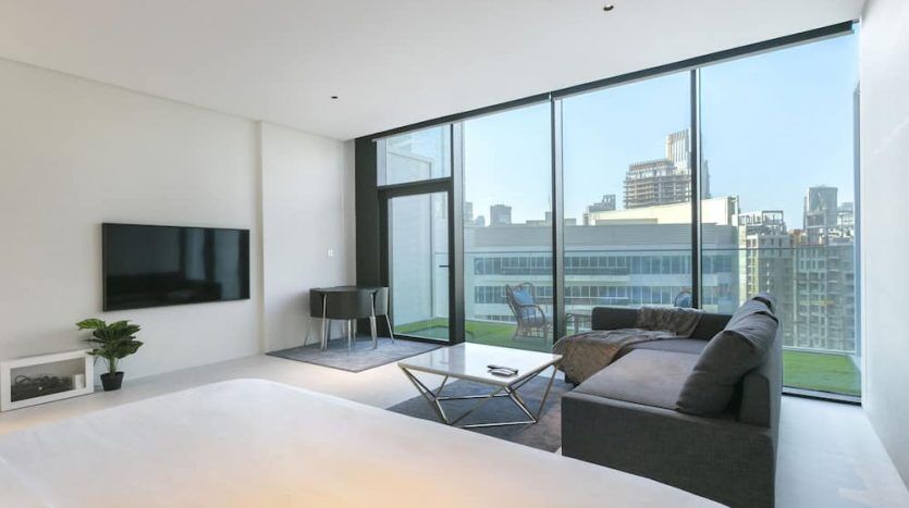 Salon d'appartement moderne à Marquise Square, Dubaï avec de grandes fenêtres offrant une vue sur la ville, comprenant un canapé gris foncé, une table basse, une télévision montée et une décoration minimaliste.
