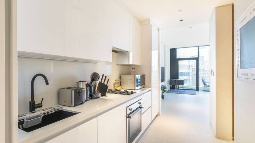 Cuisine moderne avec armoires blanches, comptoirs noirs et appareils électroménagers en acier inoxydable. Une cafetière, un grille-pain et d'autres petits appareils électroménagers sont visibles, avec un salon lumineux et naturellement éclairé en arrière-plan à Mar