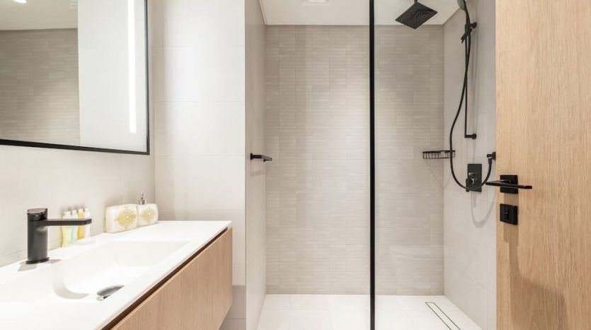 Salle de bains moderne dotée d'un comptoir blanc élégant avec un lavabo, un miroir éclairé et une douche à l'italienne spacieuse avec une pomme de douche noire et une porte vitrée sur la place Marquise, à Dubaï.