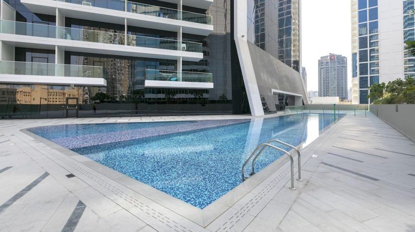 Une piscine extérieure avec des carreaux de mosaïque bleue, située sur Marquise Square Dubaï, entourée d'immeubles modernes de grande hauteur, dotée d'une terrasse propre et ouverte dans un environnement urbain.