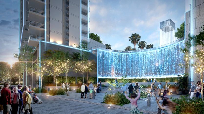 Un espace de rassemblement urbain extérieur au crépuscule à Dubaï, mettant en vedette des gens socialisant parmi des arbres illuminés et une superbe cascade de lumière bleue entre des immeubles de grande hauteur.