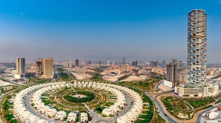 A panoramic view of a modern cityscape featuring curved residential developments and a high-rise tower in Dubai. Suburban development and construction sites stretch to the horizon under clear skies.