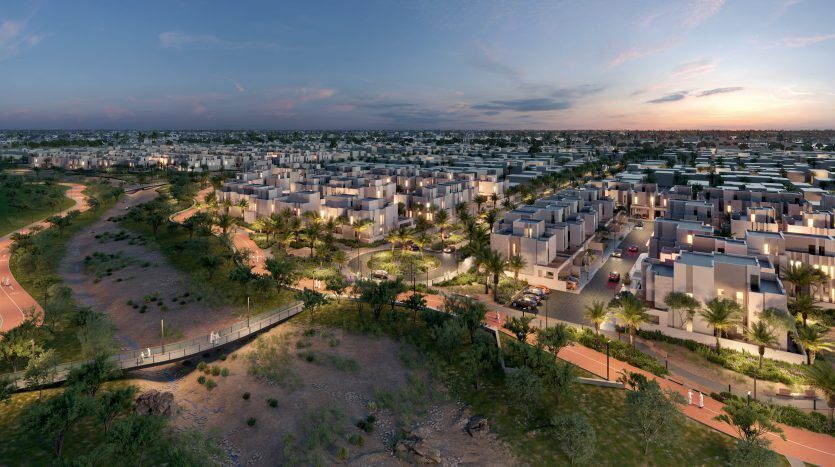 Vue aérienne d'un quartier résidentiel moderne de Dubaï avec des maisons cubiques uniformes, éclairées par un coucher de soleil, avec des sentiers sinueux et une verdure luxuriante le long d'une rivière avec un pont.