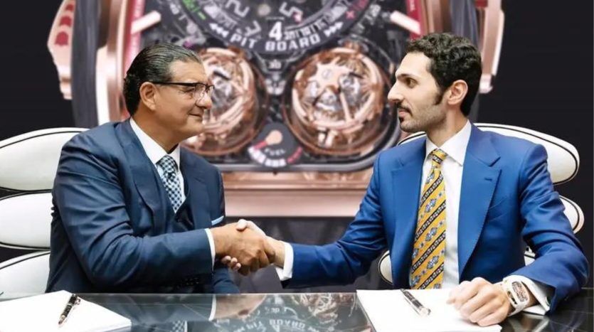 Two men in business suits shake hands around a table in a Dubai real estate office, with a large, intricate watch pattern superimposed in the background.