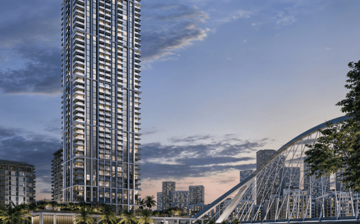 Un paysage urbain au crépuscule avec un grand immeuble moderne au premier plan, entouré de petits bâtiments. Un pont incurvé s'arche gracieusement au-dessus d'un plan d'eau à droite, mettant en valeur la beauté sereine du port de Dubai Creek près d'Emaar. Le ciel est rempli de nuages doux et d’un soupçon de soleil couchant.