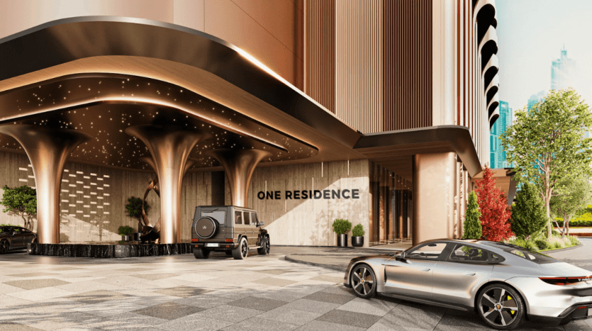 Une entrée luxueuse dans un immeuble résidentiel haut de gamme nommé « One Residences ». La scène présente une architecture contemporaine avec des colonnes épurées et un éclairage élégant. Un SUV noir et une voiture de sport argentée sont garés dans l’allée entourée d’une verdure luxuriante.