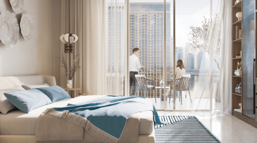Une chambre moderne avec un grand lit avec une literie bleue et beige. Des baies vitrées mènent à un balcon à Marina Cove, où deux personnes s'assoient à une table, avec vue sur le paysage urbain et le front de mer. La chambre est décorée de couleurs claires et de plantes.