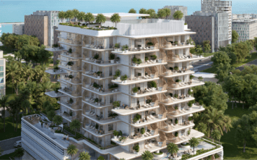 Un immeuble d'appartements moderne à plusieurs étages entouré d'arbres et doté de grands balcons verdoyants se dresse fièrement près de Sunset Bay. La structure offre un aperçu de l'océan en arrière-plan, en harmonie avec les autres bâtiments voisins le long du front de mer tranquille.