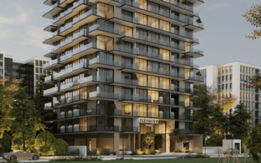 Immeuble résidentiel moderne de plusieurs étages avec de larges balcons vitrés, entouré d'arbres et de verdure, situé dans le parc Arlington de Dubaï. L'intérieur est baigné de lumière chaleureuse, quelques voitures sont garées devant et les bâtiments adjacents sont visibles en arrière-plan.