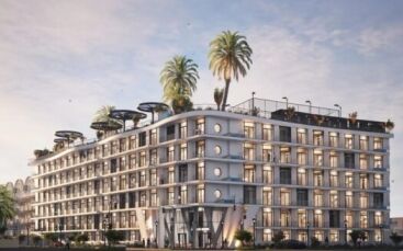 Bâtiment moderne de plusieurs étages doté de grandes fenêtres et de balcons, Avana Residences par Deca Properties à Dubaï se caractérise par des palmiers sur les toits, une végétation luxuriante et des espaces de détente extérieurs, le tout sur fond de ciel bleu vif avec quelques nuages.