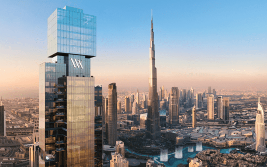 Un grand gratte-ciel moderne portant le logo "Waldorf Astoria" se dresse au premier plan, surplombant un paysage urbain composé du Burj Khalifa et des bâtiments environnants de Dubaï, sous un ciel clair au coucher du soleil.