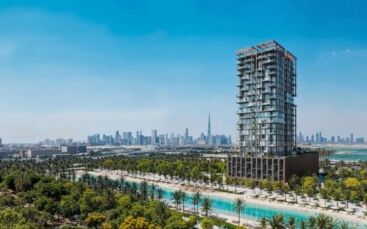 Une tour moderne, Wynwood Horizon de Imtiaz, se dresse près d'un canal bordé de palmiers, avec la ligne d'horizon de la ville et le Burj Khalifa visibles en arrière-plan sous un ciel bleu vif.