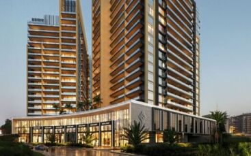 Les tours modernes Butterfly Towers Arjan, avec leurs grands balcons et leurs baies vitrées, sont illuminées le soir. Une zone commerciale de faible hauteur avec des magasins et des palmiers se trouve devant, reflétant les lumières de la ville sur la chaussée mouillée.