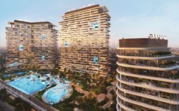 Un complexe moderne d'immeubles de grande hauteur avec des balcons uniques en quinconce, doté d'une grande piscine centrale au milieu des palmiers. Situés dans le quartier Damac de Dubaï, les bâtiments arborent le logo "DAMAC" et surplombent un paysage urbain au coucher du soleil.