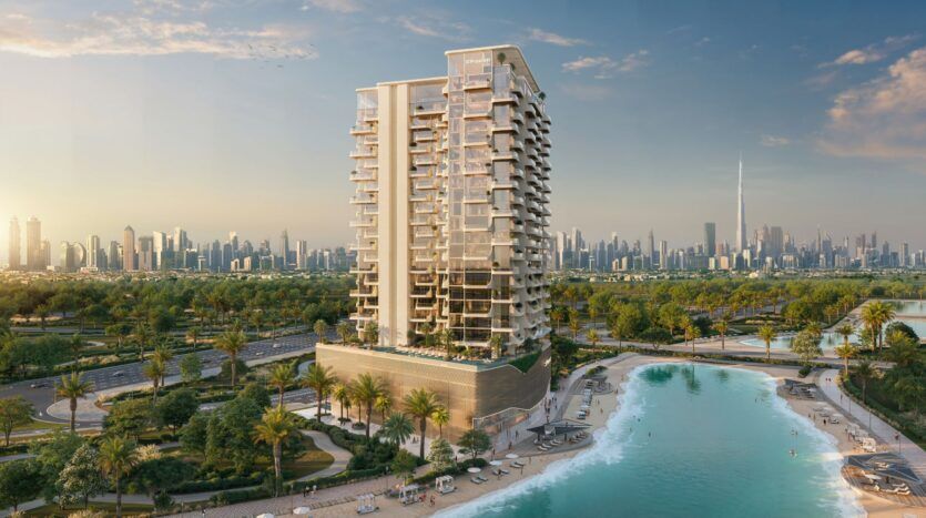 Le Caden est un immeuble résidentiel moderne de grande hauteur situé au bord d'un lagon turquoise avec des plages de sable, entouré de palmiers. La ligne d'horizon de Dubaï, y compris le Burj Khalifa, est visible à l'arrière-plan sous un ciel clair.