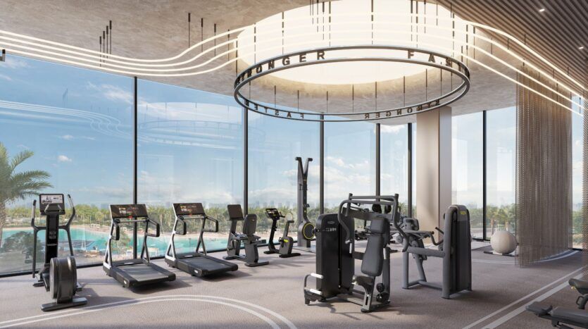 Le Caden dispose d'une salle de sport moderne équipée de tapis roulants, d'elliptiques et d'appareils de musculation, avec des fenêtres allant du sol au plafond et donnant sur une piscine et des palmiers. Un remarquable luminaire circulaire est suspendu au plafond.