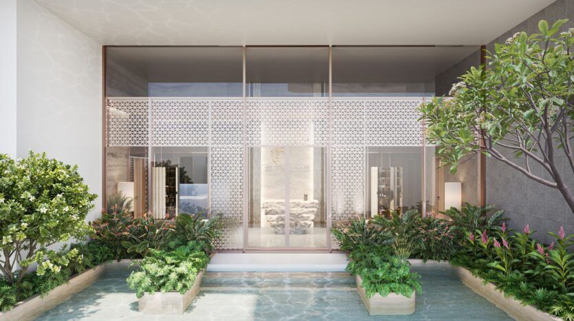 L'entrée du bâtiment moderne du Caden comporte de grandes portes et fenêtres en verre, entourées de plantes et d'arbres verdoyants. Un plan d'eau peu profond avec des jardinières se trouve au premier plan, reflétant la lumière et la verdure.