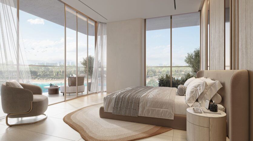 La chambre à coucher moderne et ensoleillée du Caden est dotée de fenêtres allant du sol au plafond, d'une palette neutre, d'un grand lit et d'un fauteuil confortable, le tout encadrant des vues sereines sur la piscine, la verdure luxuriante et le ciel à travers d'élégants rideaux diaphanes.