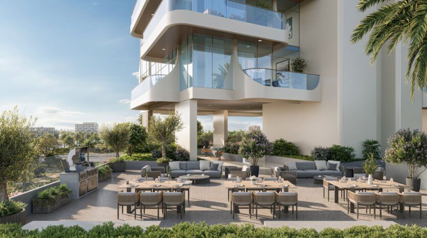 Salle à manger en plein air avec plusieurs tables et chaises installées sur un patio entouré de verdure. L'espace est surplombé par le Caden, un bâtiment moderne aux balcons de verre et à l'architecture courbée sous un ciel dégagé.