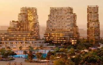 Au coucher du soleil, des gratte-ciel modernes de Hado avec des façades en verre et de la verdure se dressent parmi les arbres, reflétant une lumière chaude. Une piscine et des espaces extérieurs aménagés sont visibles au premier plan, créant une atmosphère urbaine luxuriante.