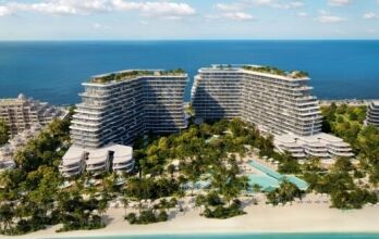 Vue aérienne des bâtiments modernes et incurvés de Bella by Passo, situés en bord de mer et entourés d'une végétation luxuriante, de piscines et d'espaces de détente. Ils sont parfaitement situés le long d'une plage de sable et d'un océan turquoise, sous un ciel bleu parsemé de nuages.