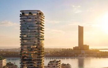 Une tour moderne, Le Château, aux balcons incurvés et à la végétation luxuriante, se dresse au bord de l'eau au coucher du soleil, tandis qu'une autre grande tour s'élève à l'arrière-plan au-dessus des eaux calmes reflétant la chaleur du ciel.