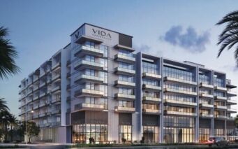 Immeuble moderne de plusieurs étages avec balcons en verre et grandes fenêtres, portant l'inscription "VIDA RESIDENCES" au sommet. Des palmiers et des aménagements paysagers encadrent la scène, dans un ciel crépusculaire - une vue emblématique de Vida Residences Saadiyat Island.