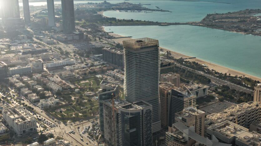 Vue aérienne d'une ville côtière où se trouve Vida Residences Saadiyat Island, avec des immeubles de grande hauteur, des routes et des espaces verts au bord d'une mer turquoise. En arrière-plan, des plages et une étendue de terre s'avancent dans l'eau.