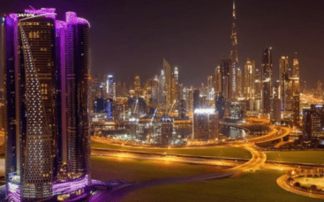 Vue nocturne d'un paysage urbain bien éclairé avec des gratte-ciel modernes, dont une tour avec un éclairage violet, et des routes courbes et éclairées. La Burj Khalifa et les Damac Towers Paramount sont visibles
