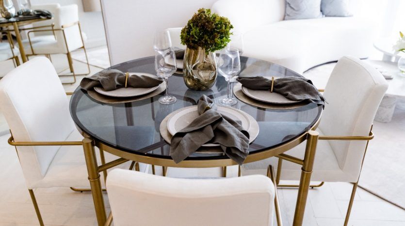 Configuration de salle à manger élégante comprenant une table ronde en verre avec des bordures dorées, accompagnée de quatre chaises blanches. La table est soigneusement dressée avec des assiettes, des verres, des serviettes et une petite pièce maîtresse en forme de plante verte d'Oxford.