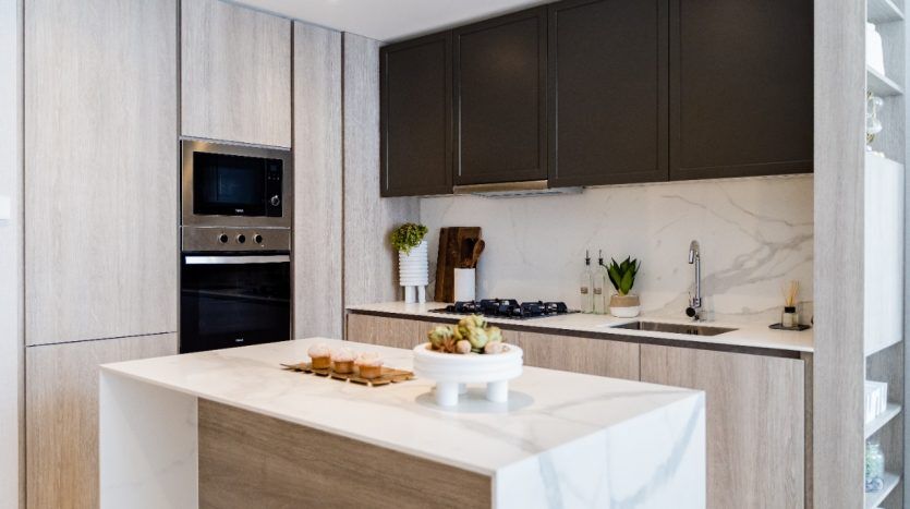 Intérieur de cuisine moderne avec armoires en bois élégantes, comptoirs en marbre et appareils électroménagers intégrés en acier inoxydable. Un îlot central abrite un présentoir à gâteaux Oxford 212 avec pâtisseries.