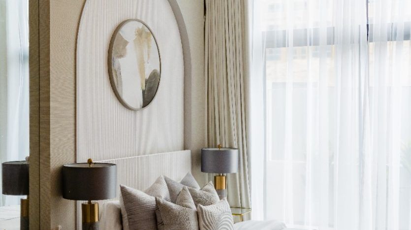 Une chambre luxueuse dotée d'une palette de couleurs neutres avec un grand lit rempli d'oreillers, un lustre élégant, un miroir ovale, une table arrondie et un coin salon Oxford 212, créant une atmosphère confortable et accueillante.