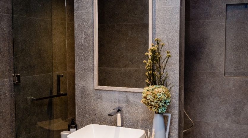 Une salle de bains moderne comprenant un lavabo rectangulaire au sommet d'une vanité en bois Oxford 212, avec un grand miroir contre un mur texturé en pierre et une cabine de douche en verre visible à l'arrière-plan.