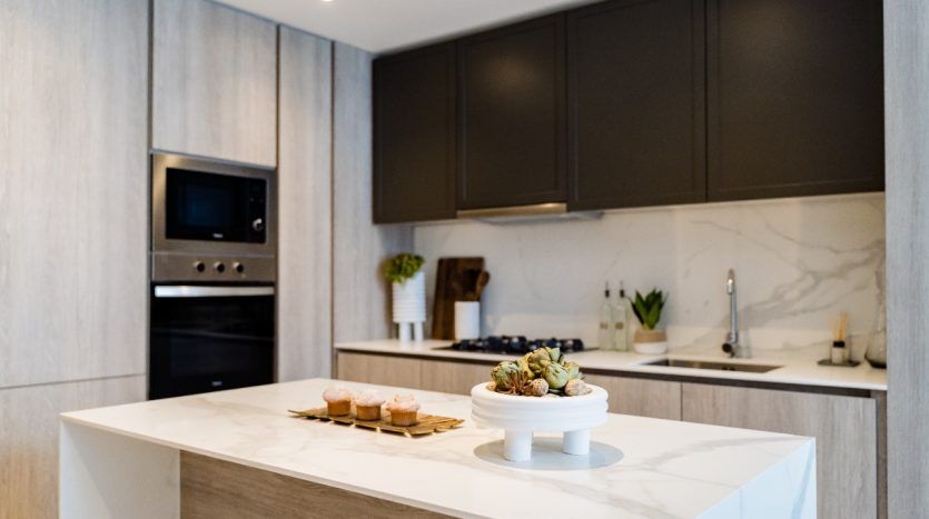 Cuisine moderne avec armoires supérieures sombres et armoires inférieures en bois clair, îlot de comptoir en marbre blanc, appareils électroménagers intégrés en acier inoxydable et plantes décoratives Oxford 212.