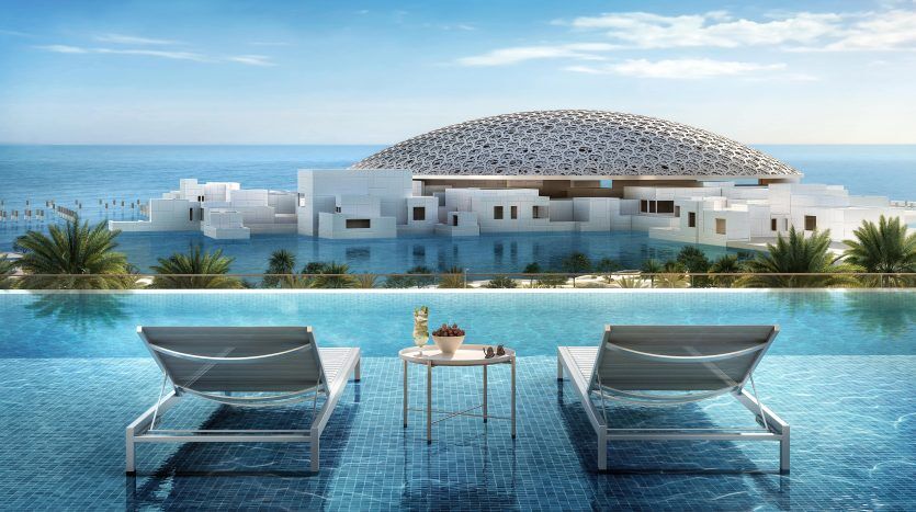 Un rendu architectural d'un bâtiment moderne surmonté d'un dôme à Dubaï au bord de la mer, vu depuis une piscine à débordement ornée de chaises longues et d'une petite table sous un ciel bleu clair.