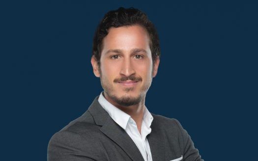 Portrait professionnel d'un homme aux cheveux noirs courts et à la barbe taillée, portant un blazer gris sur une chemise blanche, sur fond d'appartement à Dubaï.