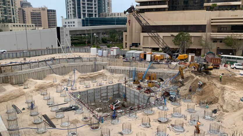 Un chantier de construction animé avec des ouvriers à Dubaï, entouré d'immeubles de grande hauteur. Excavation et piliers structurels en cours, avec une grue et divers véhicules de construction visibles.
