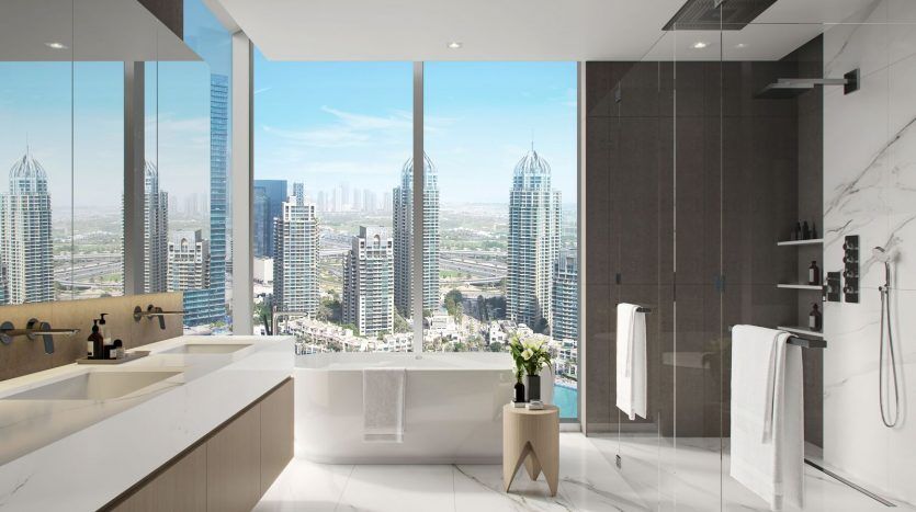 Une salle de bains luxueuse avec de grandes fenêtres offrant une vue sur les toits de la ville, comprenant une baignoire, une douche à l'italienne et deux vasques sur un comptoir en marbre. Idéal pour investir à Dubaï.