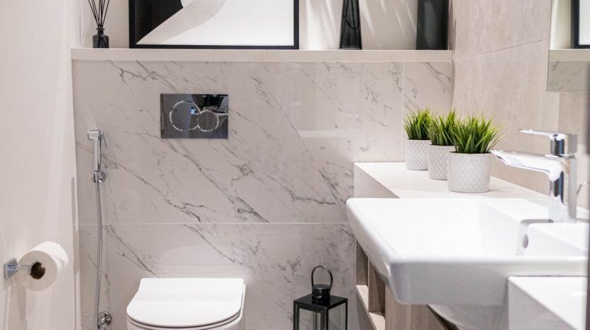 Une salle de bains moderne dans un appartement de Dubaï avec des murs en marbre élégants, comprenant des toilettes flottantes et un lavabo. Les éléments décoratifs comprennent une œuvre d'art abstraite encadrée, des plantes vertes et des accessoires minimalistes.