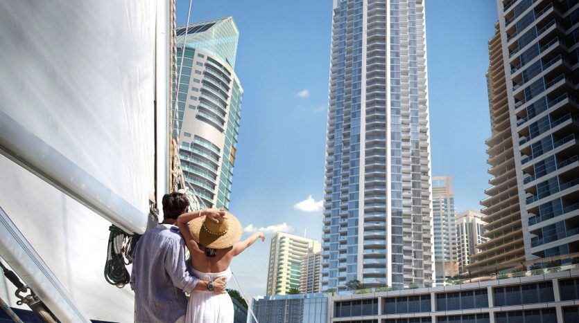 Un couple s'embrasse sur un yacht, surplombant les toits d'une ville moderne avec les imposants gratte-ciel de Dubaï et un ciel bleu clair. La femme tient un chapeau de soleil, indiquant une journée ensoleillée.