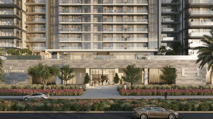 L'image montre la Ellington House à Dubai Hills, un immeuble résidentiel moderne avec une élégante façade en verre. Des palmiers et un aménagement paysager luxuriant ornent la façade, tandis qu'un chemin bordé de verdure mène à l'entrée. Deux voitures sont garées dans la rue à proximité, complétant cette oasis urbaine sereine.
